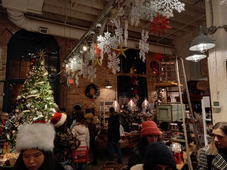 The inside of a shop has red and white snowflakes strung across the ceiling, a Christmas tree in the corner, and people walking around, looking at things to buy.