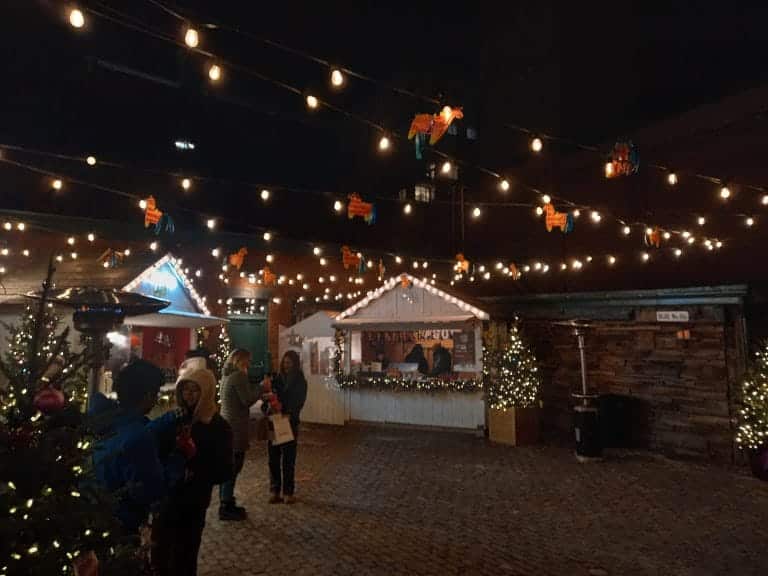 Twinkle lights with lama pinatas are strung in the sky between two little cabins with people walking between them at night.