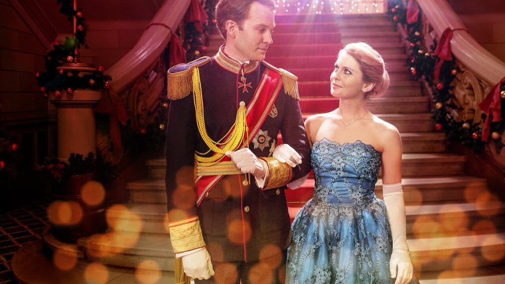 A young prince and a princess arm in arm while looking at each other and standing in front of a grand staircase.