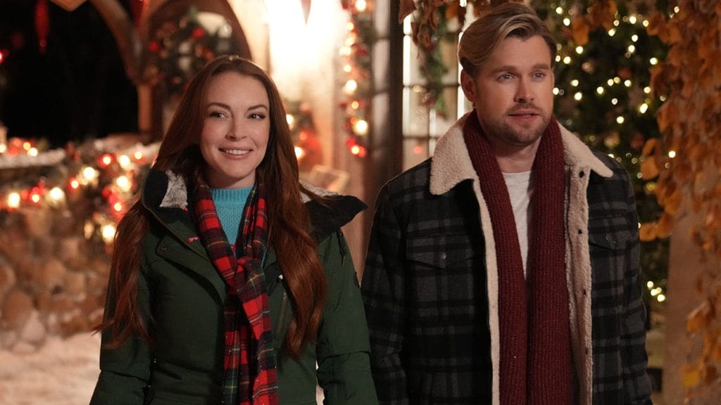 A man and a woman smiling while standing beside each other outside in front of a display of Christmas lights. 