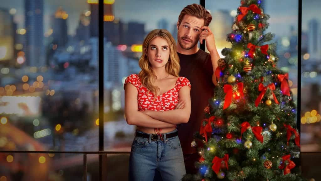 A man and woman standing next to each other beside a multi-coloured Christmas tree. The woman has her arms crossed. And, as a result of this being a romantic comedy, they are also making funny faces.