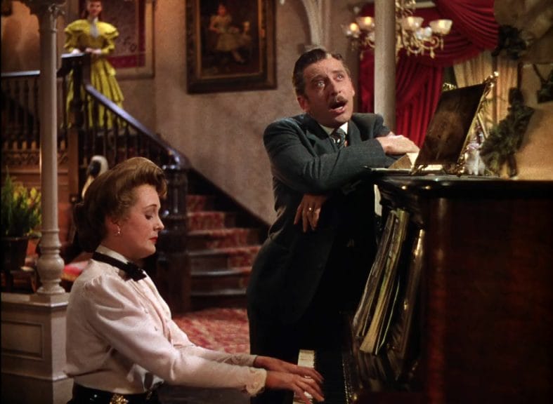 A woman plays the piano while a man leans against it and sings along.