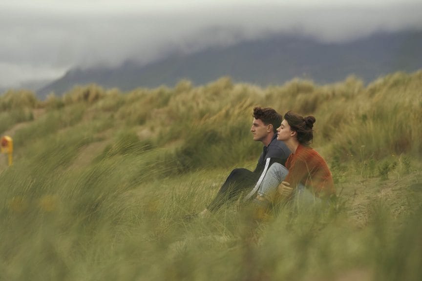 Marianne and Connell sit side by side in a hazy field, watching the horizon and surrounded by tall grass.