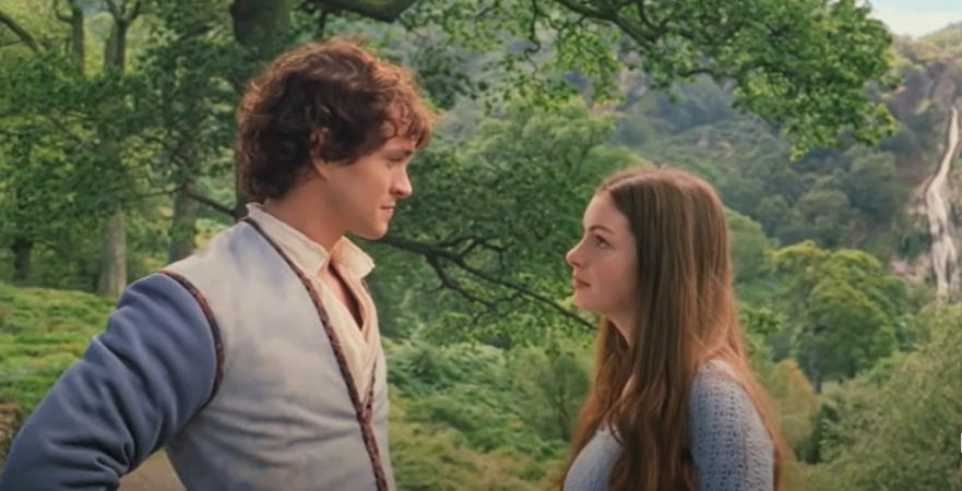 Ella, a woman with dark hair looks up at Charmont, a man with curly brown hair wearing a medieval tunic. They are both in the woods