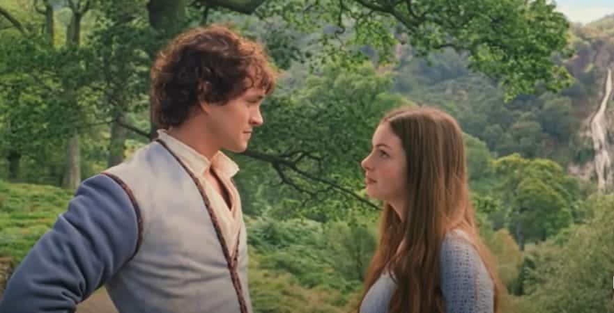 Ella, a woman with dark hair looks up at Charmont, a man with curly brown hair wearing a medieval tunic. They are both in the woods