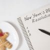 Notebook page titled "New Year's 2024 Resolutions" with a list numbered 1 to 3. A pen is sitting on the notebook and a gingerbread man is sitting next to it.