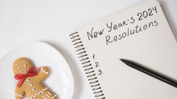 Notebook page titled "New Year's 2024 Resolutions" with a list numbered 1 to 3. A pen is sitting on the notebook and a gingerbread man is sitting next to it.