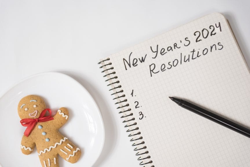 Notebook page titled "New Year's 2024 Resolutions" with a list numbered 1 to 3. A pen is sitting on the notebook and a gingerbread man is sitting next to it.