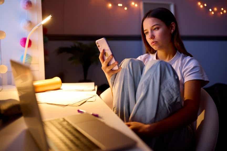 Worried teenage girl sat scrolling on her phone in her bedroom with her laptop on the table.
