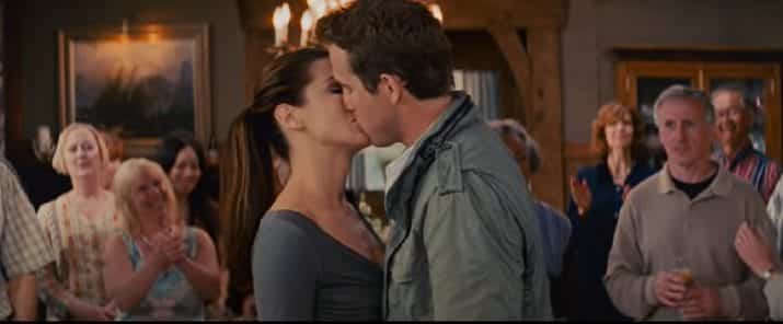 Andrew, a man with short brown hair and a grey-green jacket kisses Margaret, a woman with dark hair in a ponytail wearing grey. They are surrounded by people in a chalet room.