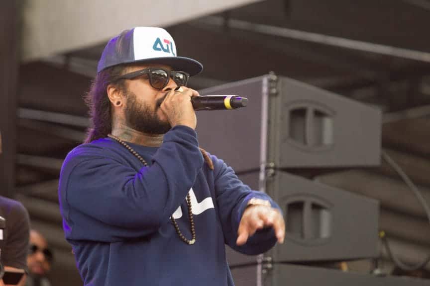 Man wearing a baseball cap and long sleeve shirt stands on stage holding a microphone