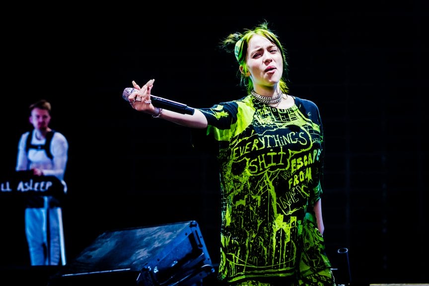 Singer-songwriter Billie Eilish, dressed in all green, holds her microphone.
