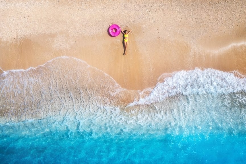 Aerial view of a woman with a pink tube float lying on the beach in the sand as waves come up on the shore. 