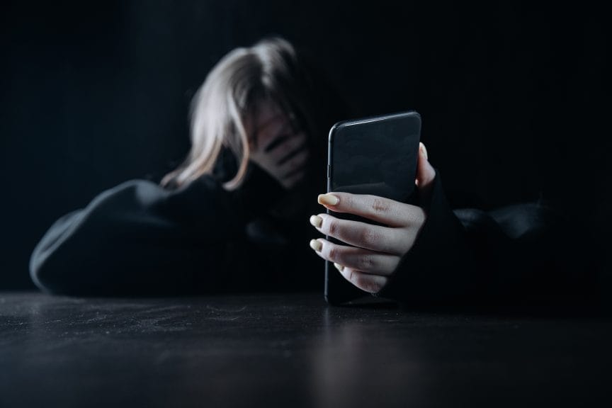 Woman has phone in one hand, covering her face with the other. She seems tired or upset, leaning forward on a table in a dark room.