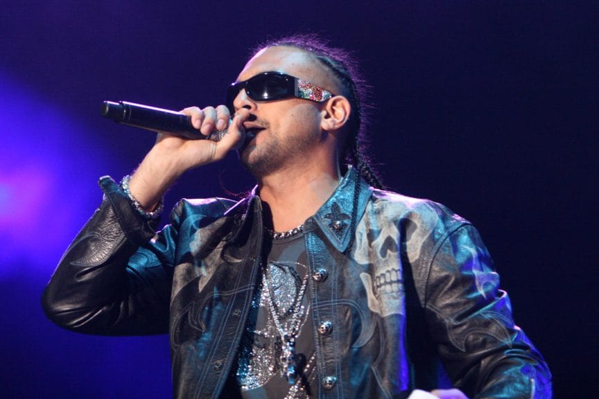 Jamaican man in sunglasses, leather jacket and chains holds a microphone on stage