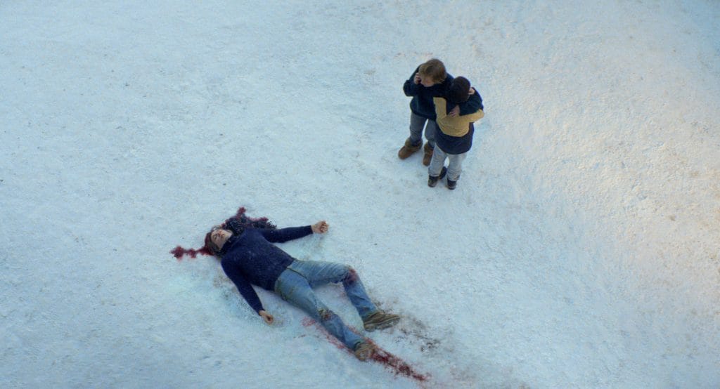 A man lays dead in the snow with his wife and son standing over him. 