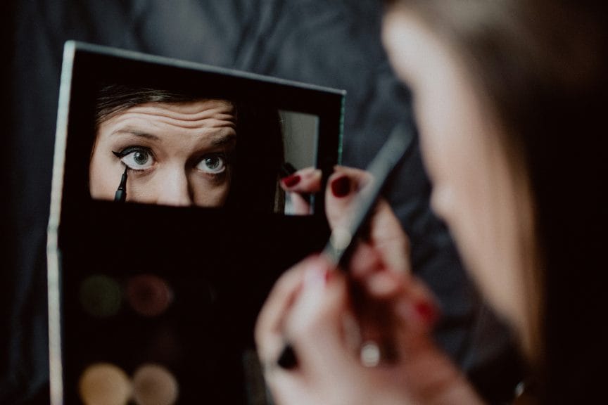 Middle-aged woman holding a palette with a mirror inside while putting on black eyeliner.