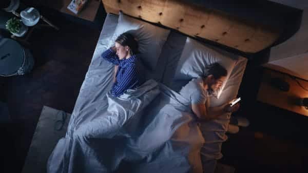 Man and woman are laying in bed on their sides facing opposite directions. The man is scrolling on his phone.