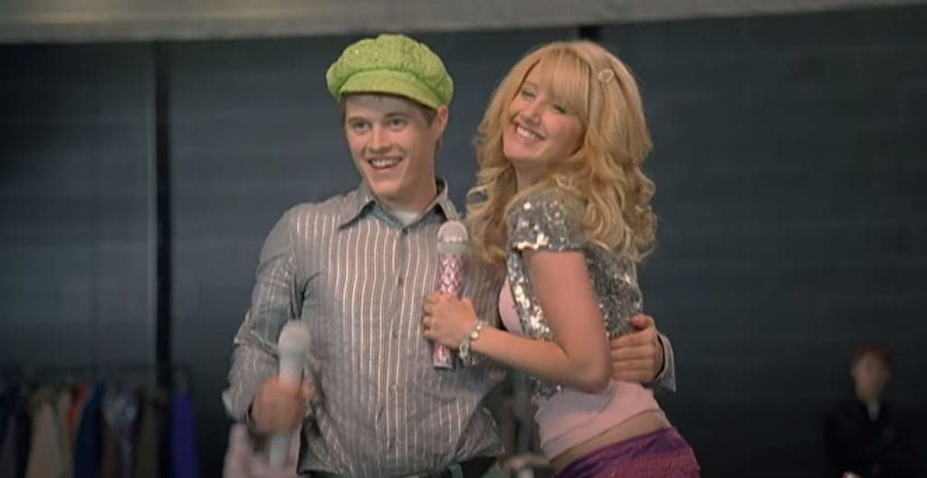 Sharpay, a smiling blonde girl holding a microphone, hugs a boy wearing a green hat