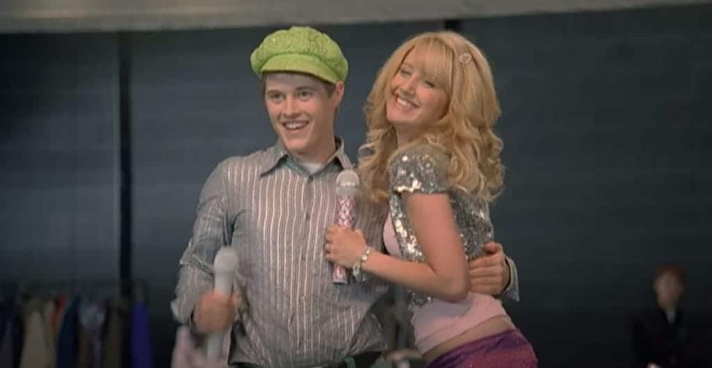 Sharpay, a smiling blonde girl holding a microphone, hugs a boy wearing a green hat