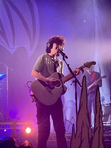 Cavetown plays a guitar, singing into a microphone against a purple background