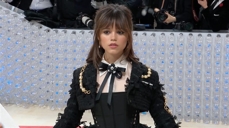 Jenna Ortega is seen at the 2023 Met Gala wearing an elegant black dress and jacket with gold chains. She is also wearing a white shirt with a black bow. 