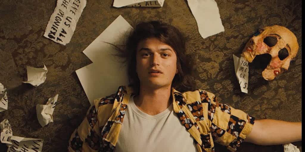 Joe Keery as Baron in the upcoming film Marmalade is seen lying on his back on the ground while he stares into space. He is wearing a yellow flannel and white tee shirt, and a bunch of blank white paper surrounds his head. 