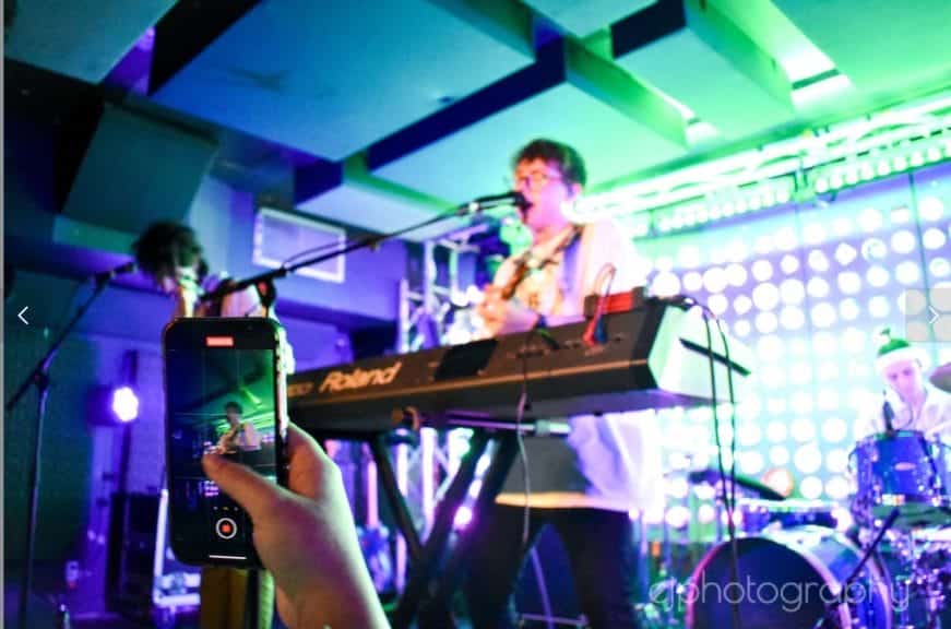 A photo of Bears in Trees performing live, with focus on someone recording it with their phone.