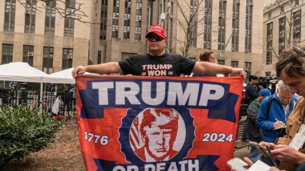trump supporter holding a homemade flag that reads "Trump or Die" with the years "1776" and "2024" on either side.