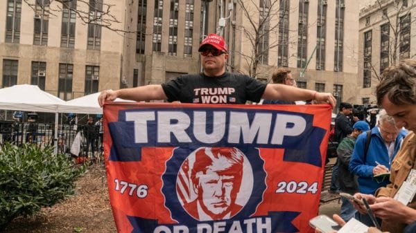 trump supporter holding a homemade flag that reads "Trump or Die" with the years "1776" and "2024" on either side.