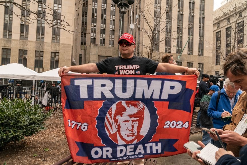 trump supporter holding a homemade flag that reads "Trump or Die" with the years "1776" and "2024" on either side.