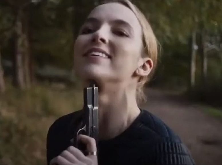 Villanelle, a woman with blonde hair and an unhinged smile holds a gun to her own head