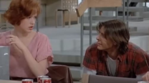 A redhead wearing a pink talk looks at a boy with black hair and a black and read check shirt. They are eating lunch in school