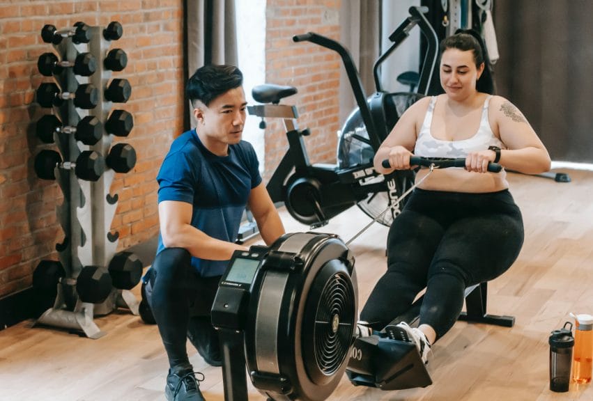 Woman working out on an exercise machine with a trainer nearby.