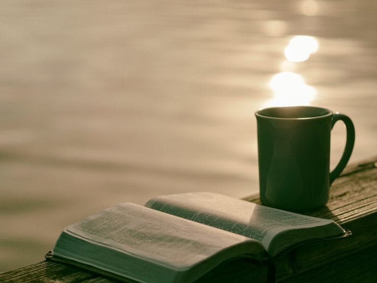 open book next to a mug of coffee or tea with a beautifully lit body of water in the background
