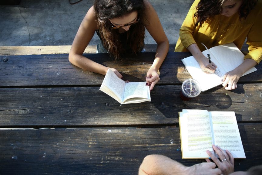 Three women having fun reading together outside at a picnic table.