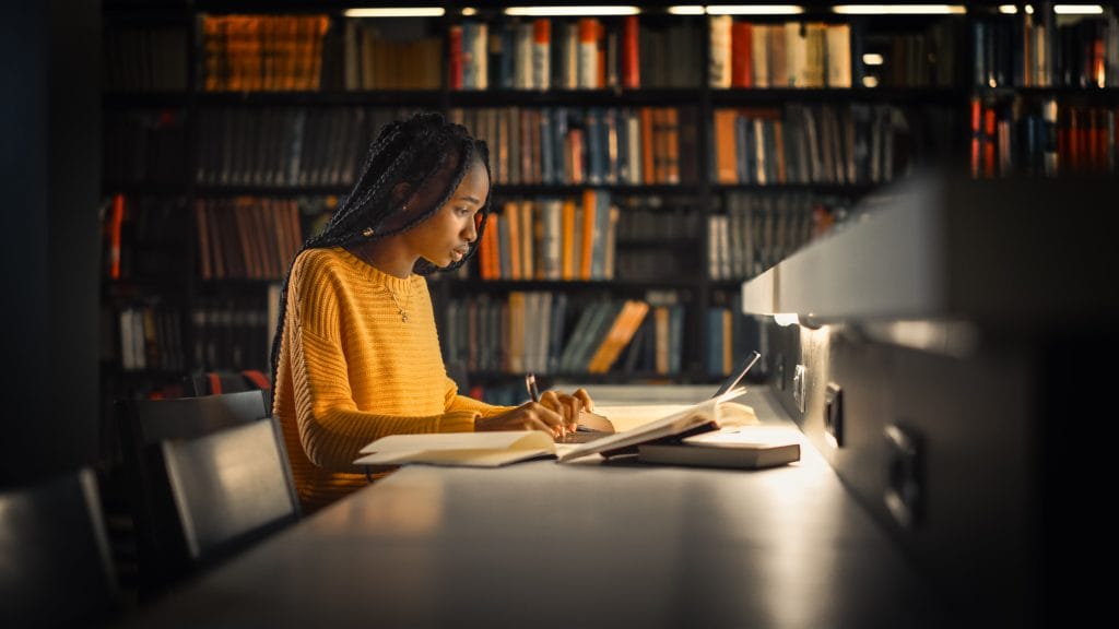 Profile of a girl in a dimly lit library working on a laptop with papers surrounding it.