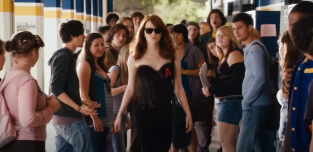 A redhead walks in a crowded highschool corridor wearing sunglasses and a black dress with the letter a on it