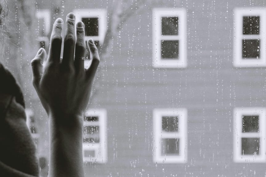 Black and white image of person's hand on the window facing representing loneliness.