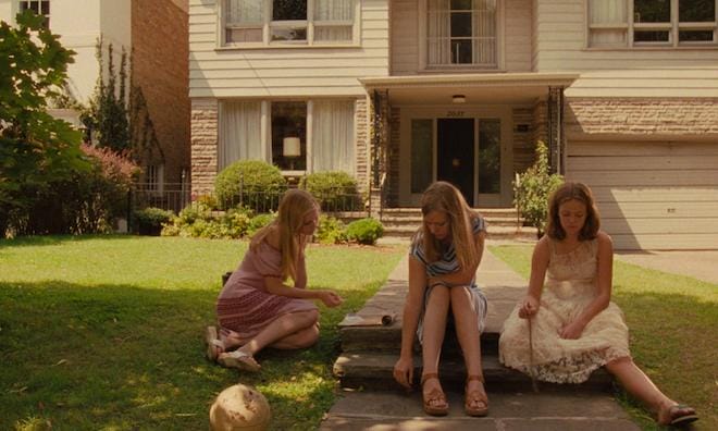 A movie still of three girls sitting on the pavement outside their surburban house.