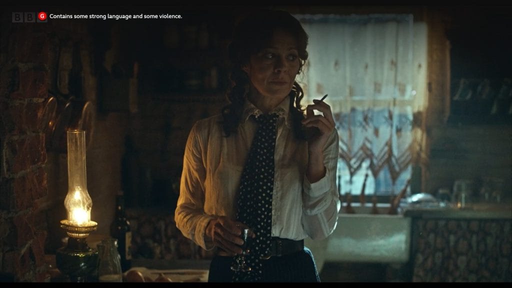 An actress with brown curly hair wears a white blouse and tie whilst smoking a cigarette