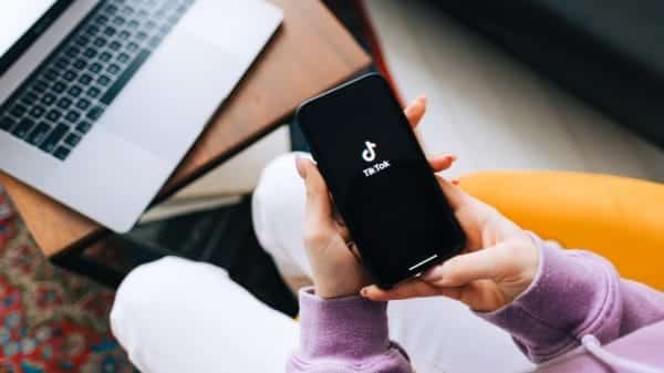 Woman holding iPhone with TikTok app logo on the screen and computer in the background