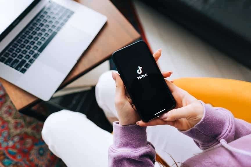 Woman holding iPhone with TikTok app logo on the screen and computer in the background