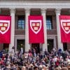 A crowd of students in front of white pillars with red flags hanging down with the Harvard emblem on them