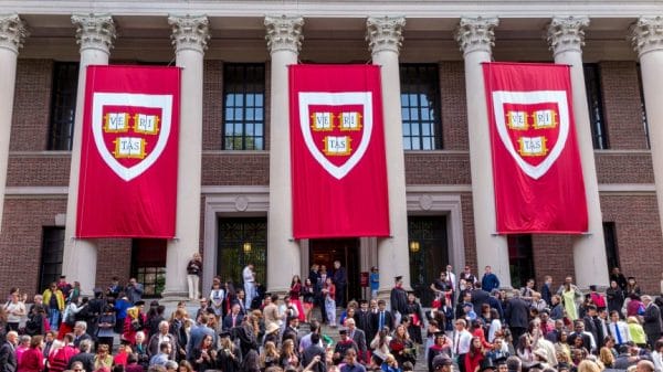A crowd of students in front of white pillars with red flags hanging down with the Harvard emblem on them