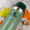 Green Stanley cup laid on concrete with fall covered leaves and acorns around it