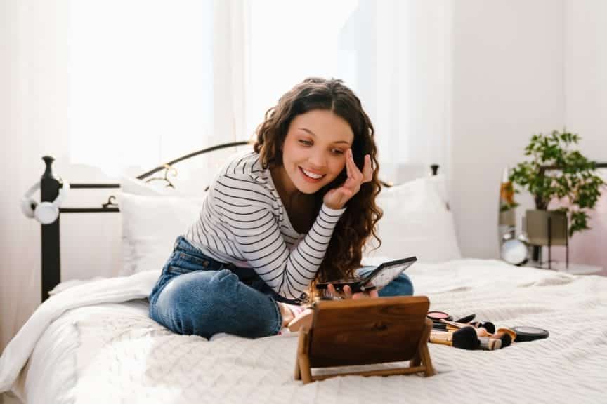 Young smiling white girl in casual wear doing makeup while sitting on bed at home