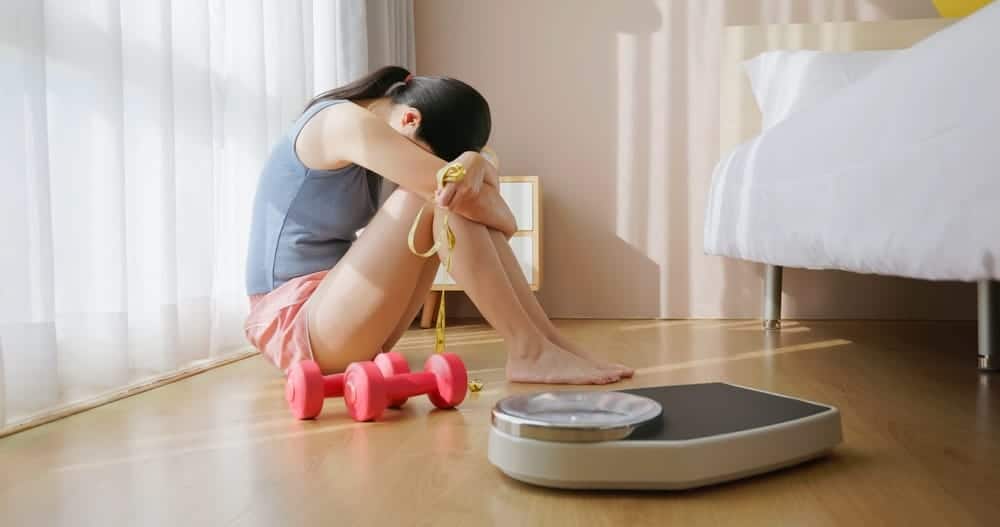 Woman is sitting on the floor, holding her head in her hands while surrounded by exercise equipment.