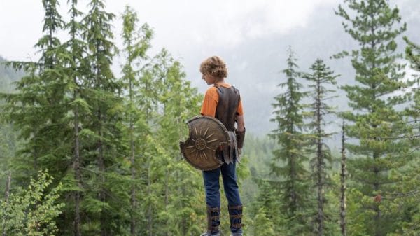 Walker Scobell as Percy Jackson, standing against a background of trees with a shield, Greek armour and an orange Camp Half-Blood t-shirt.