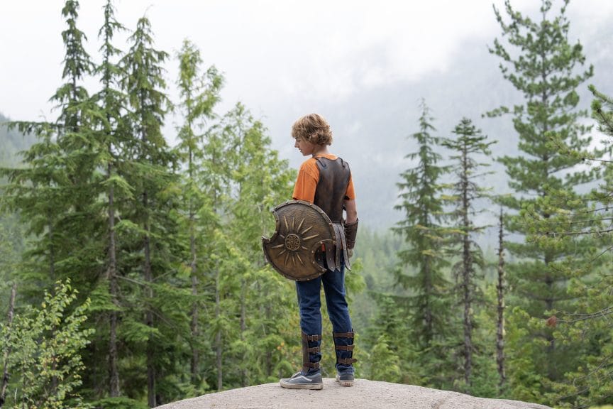 Walker Scobell as Percy Jackson, standing against a background of trees with a shield, Greek armour and an orange Camp Half-Blood t-shirt.